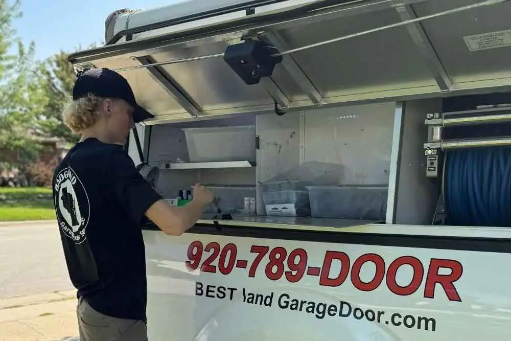 A technician from Bestland Garage Door Service in a black shirt works at an open service truck, preparing for a garage door repair in Green…| Bestland Garage Door Service – Green Bay Garage Door Repair