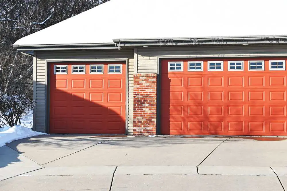 A snowy driveway leads to a two-car garage with bright orange doors featuring square windows. The scene is calm, highlighting residential charm, perfect for showcasing garage door repair services in Green Bay.| Bestland Garage Door Service – Green Bay Garage Door Repair