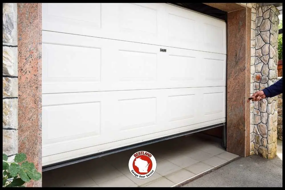 A partially open white garage door with a stone frame is shown, highlighting the need for garage door repair services in Green Bay. A person’s hand on the right side is reaching towards the door, showcasing Bestland Garage Door Service expertise.| Bestland Garage Door Service – Green Bay Garage Door Repair