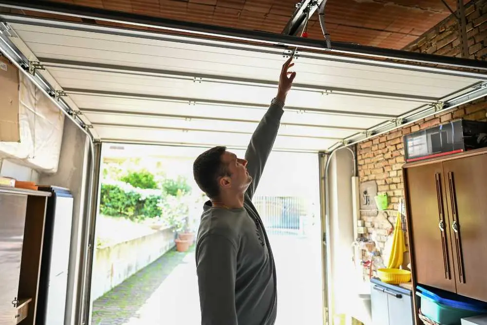 A man in a gray sweater reaches up to adjust a garage door inside a spacious garage. Sunlight filters in, highlighting garden plants outside. This scene represents professional garage door repair services in Green Bay and Appleton.| Bestland Garage Door Service – Green Bay Garage Door Repair